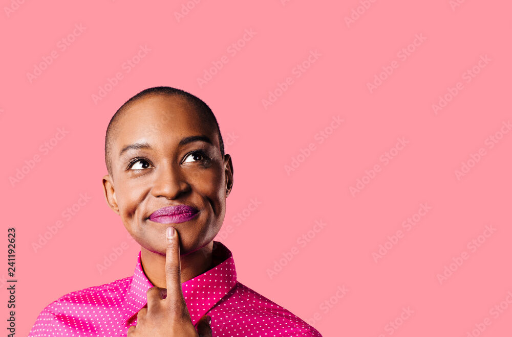 Portrait of a young woman in pink shirt with finger on mouth looking up thinking, isolated on pink studio background